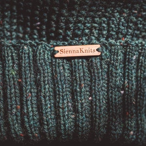Op de afbeelding: Een donkergroene gebreide beanie met een bruin leren label met de tekst "Sienna Knits".
