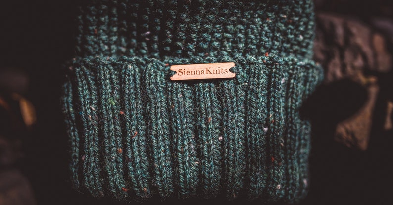 Op de afbeelding: Een donkergroene gebreide beanie met een bruin leren label met de tekst "Sienna Knits".