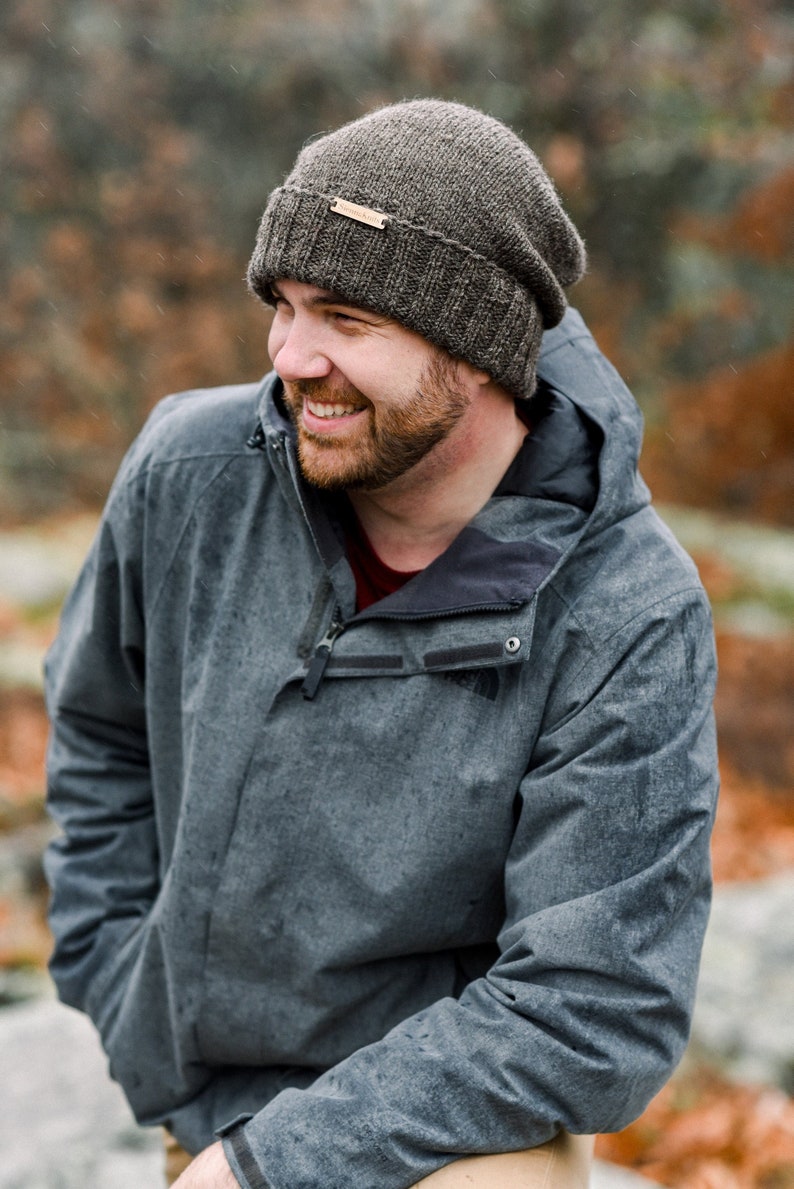 K&ouml;nnte beinhalten: Eine gestrickte graue M&uuml;tze mit einem kleinen Lederetikett wird von einer Person getragen. Die Person tr&auml;gt eine graue Kapuzenjacke. Der Hintergrund ist verschwommen mit Herbstfarben.