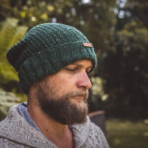Op de afbeelding: Een close-up van een persoon die een donkergroene gebreide beanie draagt met een bruin lederen label. De beanie heeft een textuurpatroon en is naar beneden getrokken over het voorhoofd van de persoon. De persoon heeft een baard en kijkt naar beneden.