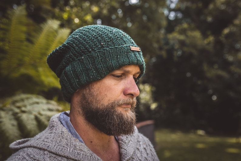 Op de afbeelding: Een close-up van een persoon die een donkergroene gebreide beanie draagt met een bruin lederen label. De beanie heeft een textuurpatroon en is naar beneden getrokken over het voorhoofd van de persoon. De persoon heeft een baard en kijkt naar beneden.