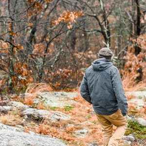 K&ouml;nnte beinhalten: Eine Person mit einer grauen Kapuzenjacke, braunen Hosen und einer Strickm&uuml;tze geht durch einen Wald. Die B&auml;ume haben kahle &Auml;ste und orangefarbene Bl&auml;tter. Der Boden ist mit Felsen und trockenem Laub bedeckt.