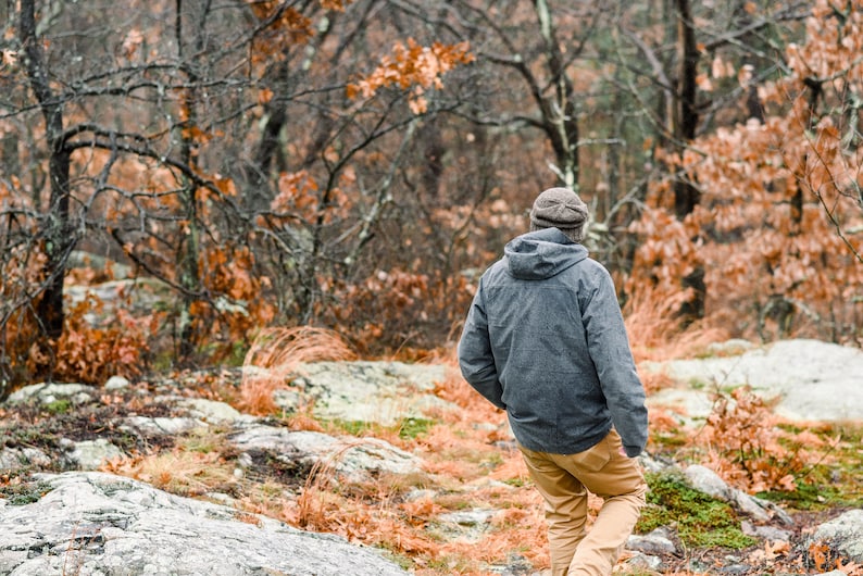 K&ouml;nnte beinhalten: Eine Person mit einer grauen Kapuzenjacke, braunen Hosen und einer Strickm&uuml;tze geht durch einen Wald. Die B&auml;ume haben kahle &Auml;ste und orangefarbene Bl&auml;tter. Der Boden ist mit Felsen und trockenem Laub bedeckt.