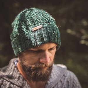 Op de afbeelding: Een close-up van een persoon die een groene gebreide beanie draagt met een bruin leren label waarop "Simple Knit" staat. De beanie is gemaakt van zacht, getextureerd garen.