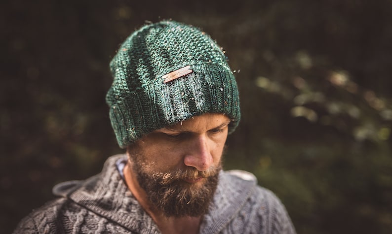 Op de afbeelding: Een close-up van een persoon die een groene gebreide beanie draagt met een bruin leren label waarop "Simple Knit" staat. De beanie is gemaakt van zacht, getextureerd garen.