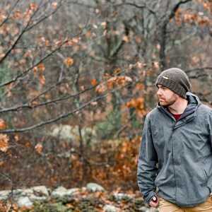 K&ouml;nnte beinhalten: Eine Person tr&auml;gt eine graue Kapuzenjacke, eine braune Strickm&uuml;tze und eine beige Hose im Freien. Der Hintergrund zeigt einen Wald mit B&auml;umen und Herbstlaub. Die Jacke hat einen Rei&szlig;verschluss und ein rotes Hemd ist darunter sichtbar.