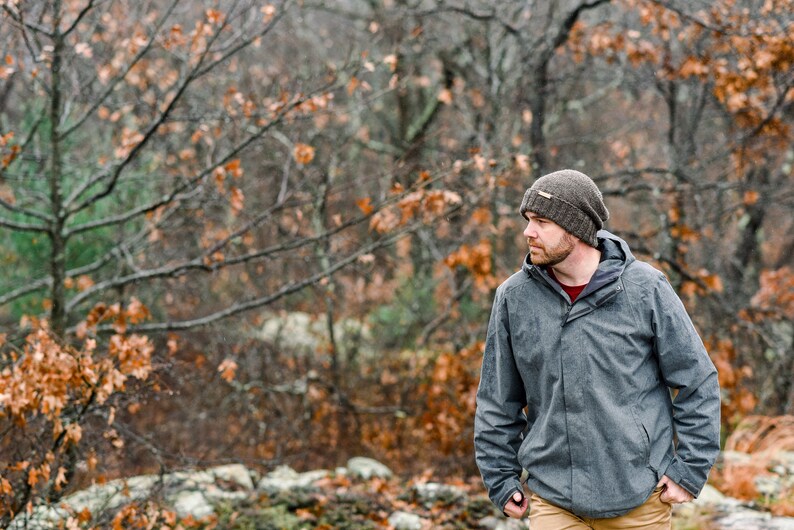 K&ouml;nnte beinhalten: Eine Person tr&auml;gt eine graue Kapuzenjacke, eine braune Strickm&uuml;tze und eine beige Hose im Freien. Der Hintergrund zeigt einen Wald mit B&auml;umen und Herbstlaub. Die Jacke hat einen Rei&szlig;verschluss und ein rotes Hemd ist darunter sichtbar.