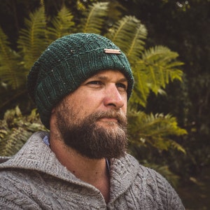 Op de afbeelding: Een man draagt ​​een groene gebreide beanie met een bruin lederen label en een grijze gebreide trui. Hij kijkt naar de zijkant en heeft een baard.