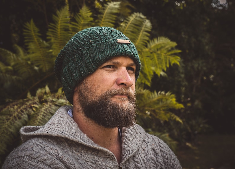 Op de afbeelding: Een man draagt ​​een groene gebreide beanie met een bruin lederen label en een grijze gebreide trui. Hij kijkt naar de zijkant en heeft een baard.