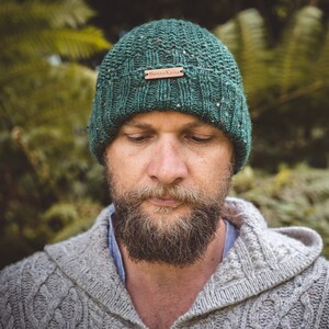 Op de afbeelding: Een man draagt ​​een groene gebreide beanie met een bruin leren label met de tekst "Sierra Kaia" en een grijze gebreide trui.