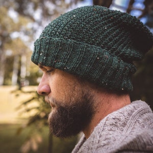 Op de afbeelding: Een man draagt ​​een groene gebreide beanie met een textuurpatroon. Hij heeft een baard en draagt ​​een lichtgrijze trui.