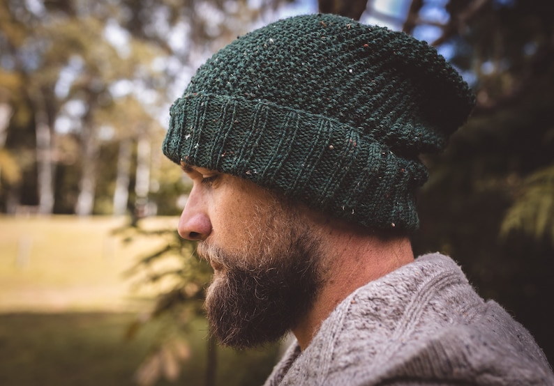 Op de afbeelding: Een man draagt ​​een groene gebreide beanie met een textuurpatroon. Hij heeft een baard en draagt ​​een lichtgrijze trui.