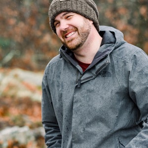 K&ouml;nnte beinhalten: Eine Person tr&auml;gt eine graue Jacke und eine braune Strickm&uuml;tze und l&auml;chelt breit. Die Jacke hat eine Kapuze und einen Rei&szlig;verschluss. Das The North Face-Logo ist auf der Brust sichtbar. Der Hintergrund ist verschwommen mit Herbstfarben.