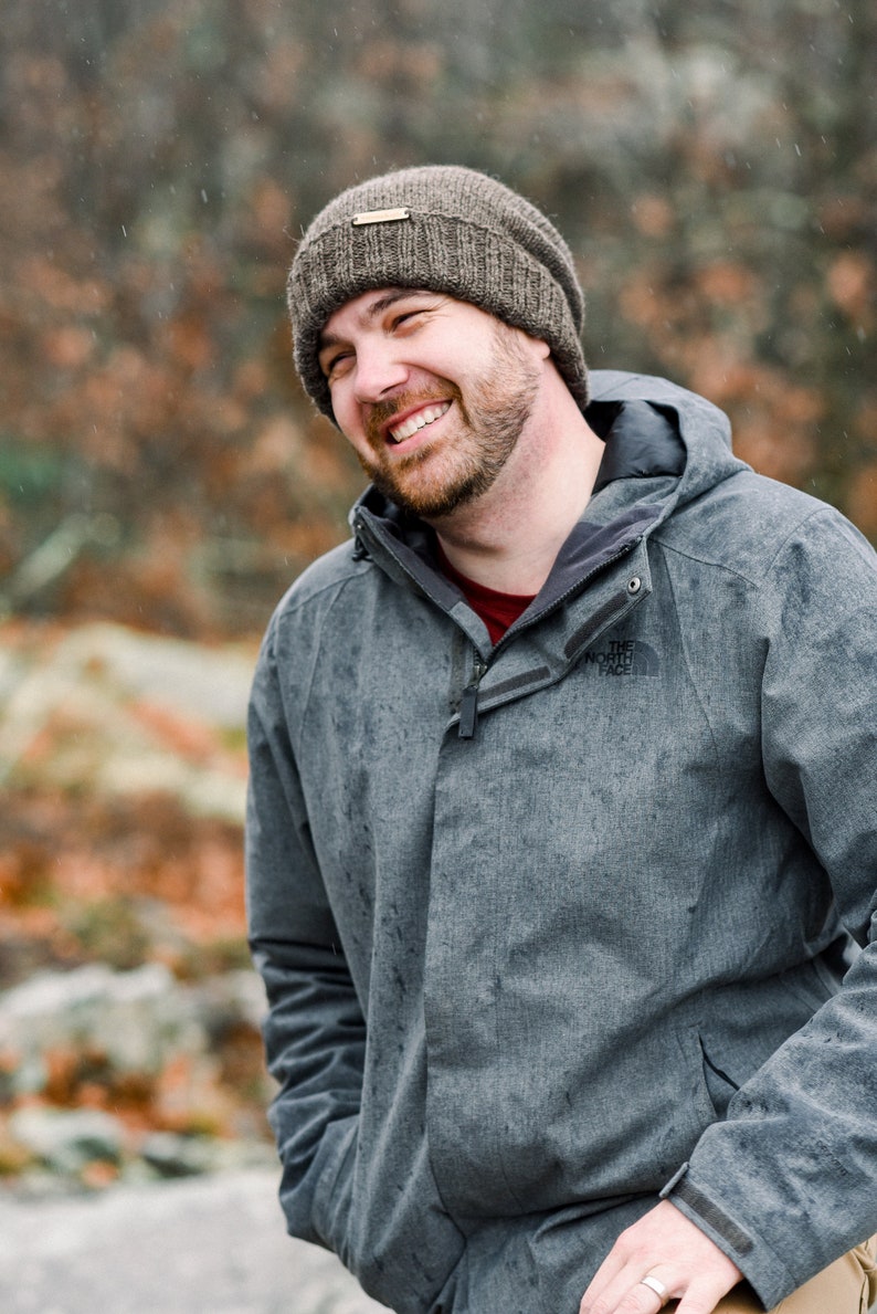 K&ouml;nnte beinhalten: Eine Person tr&auml;gt eine graue Jacke und eine braune Strickm&uuml;tze und l&auml;chelt breit. Die Jacke hat eine Kapuze und einen Rei&szlig;verschluss. Das The North Face-Logo ist auf der Brust sichtbar. Der Hintergrund ist verschwommen mit Herbstfarben.