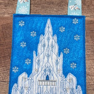 Puede incluir: Una bolsa de fieltro azul con un dise&ntilde;o de castillo bordado en blanco, detalles de purpurina plateada y un asa de cinta azul claro con motivos de copos de nieve y mu&ntilde;ecos de nieve. La bolsa tiene un tema invernal.