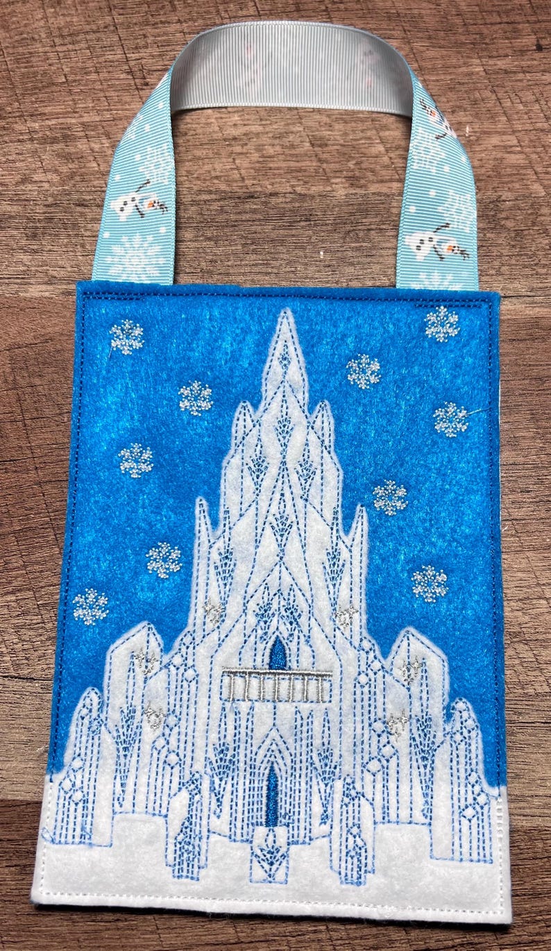 Puede incluir: Una bolsa de fieltro azul con un dise&ntilde;o de castillo bordado en blanco, detalles de purpurina plateada y un asa de cinta azul claro con motivos de copos de nieve y mu&ntilde;ecos de nieve. La bolsa tiene un tema invernal.
