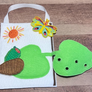 May include: A felt activity book with a white background, featuring a sun, caterpillar, and butterfly. The book has a white handle and a green leaf with holes. The caterpillar is green and brown.