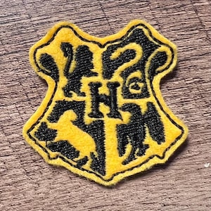 May include: A shield-shaped embroidered patch featuring a yellow background with black stitching. The design includes a central letter H surrounded by four animal silhouettes, including a lion, a snake, a badger, and an eagle, all in black.