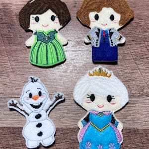 Puede incluir: Cuatro mu&ntilde;ecos de fieltro de personajes: una princesa con un vestido verde, un personaje con un traje azul, un mu&ntilde;eco de nieve y una princesa con un vestido azul con una corona dorada. Cada mu&ntilde;eco tiene detalles bordados y caras sonrientes.