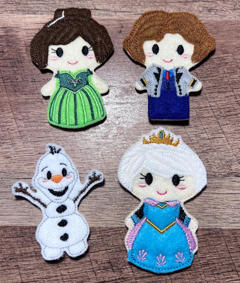 Puede incluir: Cuatro mu&ntilde;ecos de fieltro de personajes: una princesa con un vestido verde, un personaje con un traje azul, un mu&ntilde;eco de nieve y una princesa con un vestido azul con una corona dorada. Cada mu&ntilde;eco tiene detalles bordados y caras sonrientes.