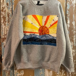 May include: A gray sweatshirt featuring a colorful, patchwork design of a sunset over blue waves. The sun is a vibrant mix of yellow, orange, and red, with rays extending outwards. The sweatshirt is displayed on a wooden hanger.
