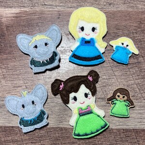 Puede incluir: Una colecci&oacute;n de mu&ntilde;ecos de fieltro de personajes, que incluyen dos elefantes y dos princesas. Los mu&ntilde;ecos presentan colores brillantes como el azul, el verde y el amarillo, con rasgos faciales y ropa detallados. Los mu&ntilde;ecos est&aacute;n dispuestos sobre una superficie de madera.