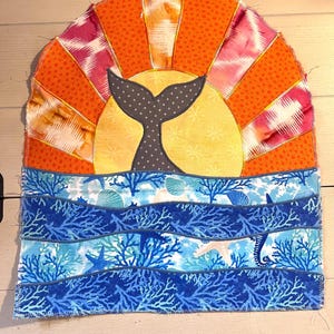 May include: A fabric quilt block featuring a dark gray whale tail silhouette centered against a yellow sun and orange sunburst background. The lower section depicts a blue ocean scene with coral, starfish, and seahorse patterns in fabric.