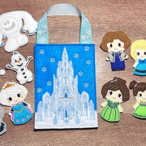 Puede incluir: Un bolso tote azul con un dise&ntilde;o de castillo blanco, rodeado de figuras de personajes de fieltro. Los personajes incluyen un mu&ntilde;eco de nieve, snowgies y princesas. El bolso tiene un asa azul claro con detalles de copos de nieve.