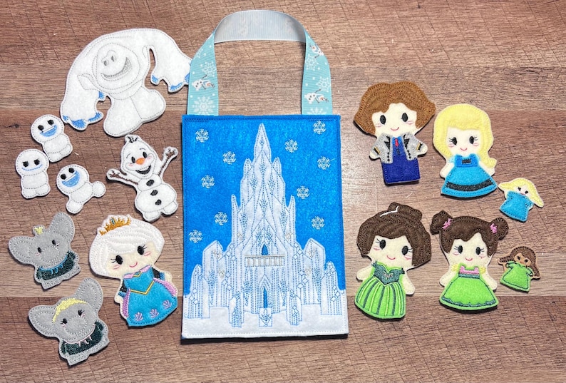 Puede incluir: Un bolso tote azul con un dise&ntilde;o de castillo blanco, rodeado de figuras de personajes de fieltro. Los personajes incluyen un mu&ntilde;eco de nieve, snowgies y princesas. El bolso tiene un asa azul claro con detalles de copos de nieve.