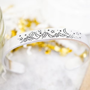 May include: A silver cuff bracelet with an engraved design of mountains, stars, and a crescent moon.