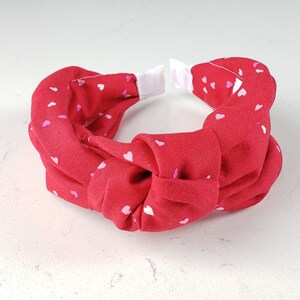 May include: Red fabric headband with a knotted design and a white polka dot pattern of small hearts.