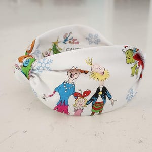 May include: A white knotted headband featuring a colorful holiday-themed print with cartoon characters, including a green creature in a red cape, a blue-clad figure, and a small child, interspersed with blue snowflakes and green Christmas trees.