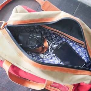 Pu&ograve; includere: Una borsa da viaggio in tela beige e rossa con cinturini e rifiniture in pelle marrone. La borsa &egrave; aperta e l'interno &egrave; foderato con un tessuto floreale blu e bianco. Una fotocamera nera e uno smartphone nero sono visibili all'interno della borsa.