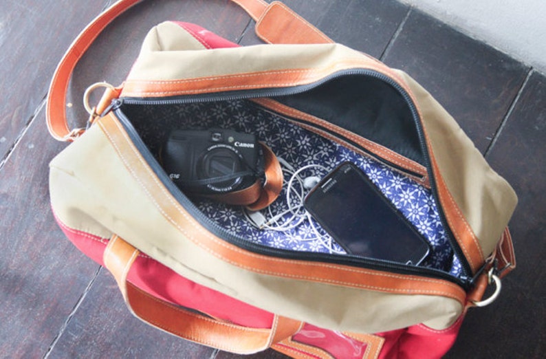 Pu&ograve; includere: Una borsa da viaggio in tela beige e rossa con cinturini e rifiniture in pelle marrone. La borsa &egrave; aperta e l'interno &egrave; foderato con un tessuto floreale blu e bianco. Una fotocamera nera e uno smartphone nero sono visibili all'interno della borsa.