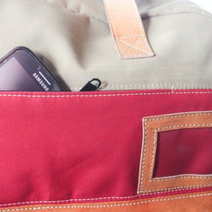 Pu&ograve; includere: Una borsa in tela beige, rossa e marrone con una tasca con cerniera e una finestra in plastica trasparente. Un telefono cellulare nero con la scritta "SAMSUNG" stampata sul retro &egrave; nella tasca.