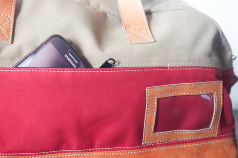 Pu&ograve; includere: Una borsa in tela beige, rossa e marrone con una tasca con cerniera e una finestra in plastica trasparente. Un telefono cellulare nero con la scritta "SAMSUNG" stampata sul retro &egrave; nella tasca.