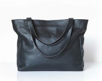 以下が含まれることがあります： 黒い革製のトートバッグ。2つの長いハンドルが付いています。バッグは長方形で、広い開口部とシンプルなデザインです。革は柔らかく、わずかに質感があり、マットな仕上がりです。日常使いに適しています。