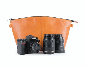 May include: A tan leather camera bag with a zip closure, a black camera body with a lens, and two additional camera lenses.