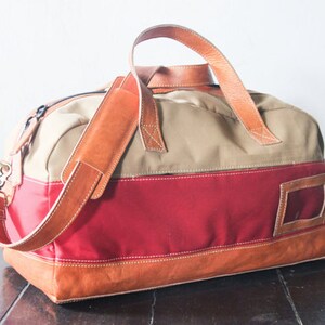 Pu&ograve; includere: Una borsa da viaggio in tela e pelle di colore beige, rosso e marrone con una lunga tracolla in pelle. La borsa ha una piccola tasca in pelle sul lato.