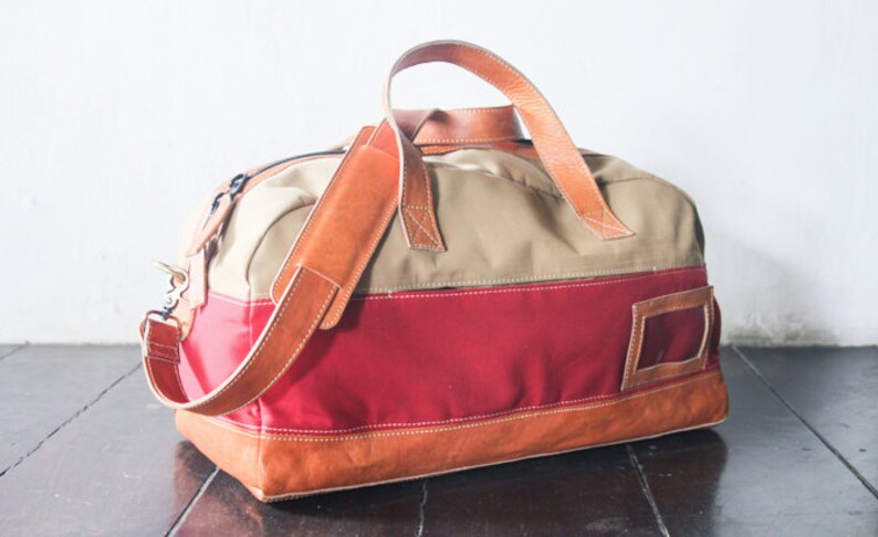 Pu&ograve; includere: Una borsa da viaggio in tela e pelle di colore beige, rosso e marrone con una lunga tracolla in pelle. La borsa ha una piccola tasca in pelle sul lato.
