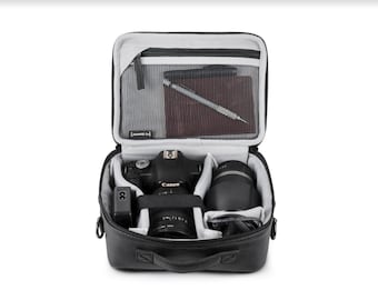May include: Black camera bag with a zipped top and a handle. The bag is open and contains a black camera, a lens, and a charger. The bag has a mesh pocket on the inside with a pen and a notebook.