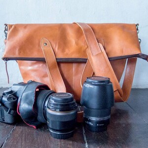 May include: A brown leather camera bag with a zipper closure and two adjustable straps. The bag is sitting on a wooden surface with a black camera and two camera lenses.