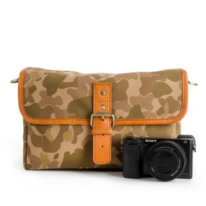 Peut inclure: Un sac à bandoulière pour appareil photo en toile à motif camouflage avec une sangle et une boucle en cuir. Un appareil photo noir avec un objectif est devant le sac.