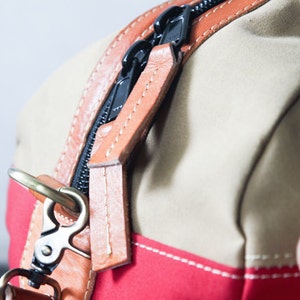 Pu&ograve; includere: Primo piano di una borsa in tela beige e rossa con un tirare zip in pelle marrone e un gancio in ottone. La cerniera &egrave; nera e le cuciture sono bianche.
