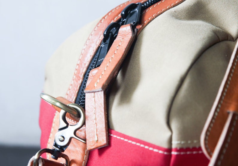 Pu&ograve; includere: Primo piano di una borsa in tela beige e rossa con un tirare zip in pelle marrone e un gancio in ottone. La cerniera &egrave; nera e le cuciture sono bianche.