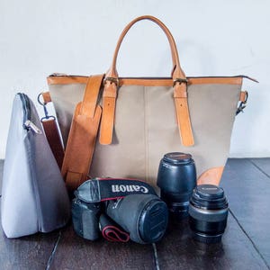 Peut inclure: Un sac à dos en cuir marron avec un corps en toile gris et une poche zippée grise. Le sac est ouvert et contient un appareil photo noir avec un objectif et deux objectifs supplémentaires.