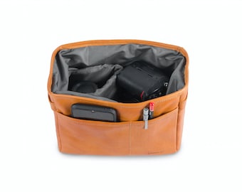 May include: A tan leather camera bag with a grey interior. The bag is open and has a camera, lens, and phone inside. The bag has a small pocket on the front with two pens.