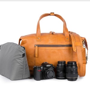 Peut inclure: Un sac à dos en cuir tanné avec un insert de caméra gris et trois objectifs d'appareil photo noirs. Le sac a une poignée supérieure et une bandoulière.