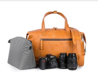 May include: A tan leather camera bag with a gray camera insert and three black camera lenses. The bag has a top handle and a shoulder strap.