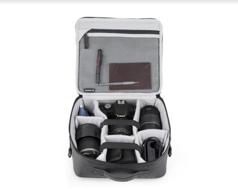 May include: A black camera bag with a zipped top compartment and a padded interior. The bag is open and contains a camera, lenses, and other accessories.